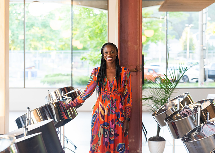 a black woman in an orange patterned dress stands in front of steel pans