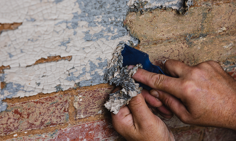 a hand scrapes layers of paint off of a brick building