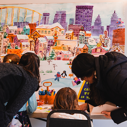 adults help a child make a drawn figure to add to a colorful mural of a city