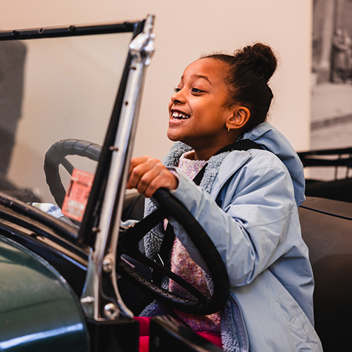 A young girl smiles as she sits behind the wheel of a vintage car