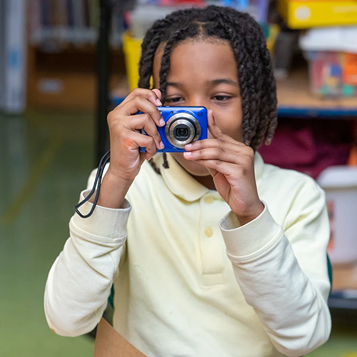 a young child with dreadlocks wears a white shirt and holds up a small blue digital camera