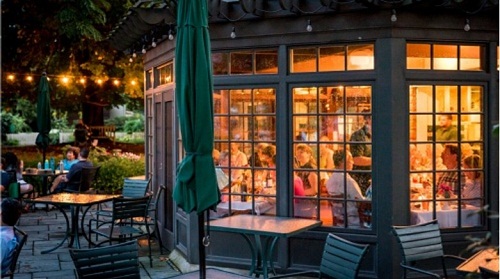 Floor-to-ceiling bow window, lit and crowded with people inside, with a patio, tables, and string lights outside