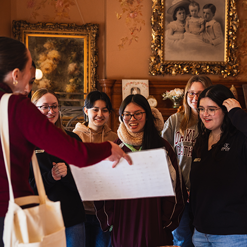a group of teenagers look at a photo being held out by a tour guide in a historic home