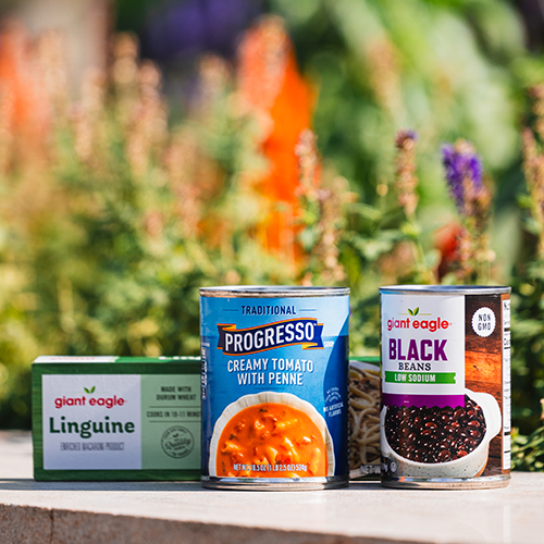 a can of soup, a can of black beans, and a box of pasta sit outside in front of blooming flowers