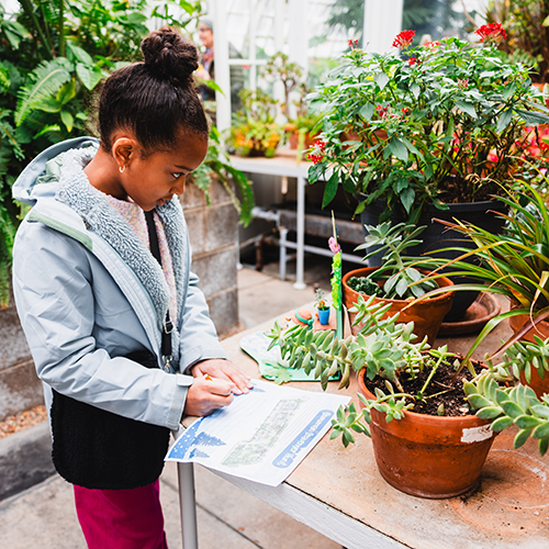 a young girl writes on a piece of paper while standing inside a greenhouse